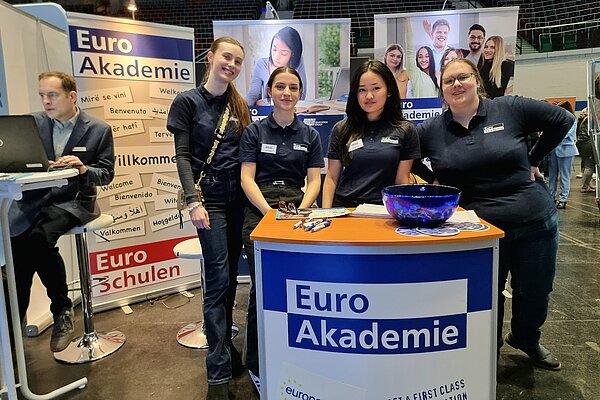 Das Bild zeigt den Messestand der Euro Akademie und der Euro-Schulen. Im Vordergrund steht das Pult mit Logo und dahinter sind vier Auszubildender im T-Shirt der Euro Akademie zu sehen, die freundlich in die Kamera blicken.