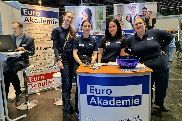 Das Bild zeigt den Messestand der Euro Akademie und der Euro-Schulen. Im Vordergrund steht das Pult mit Logo und dahinter sind vier Auszubildender im T-Shirt der Euro Akademie zu sehen, die freundlich in die Kamera blicken.