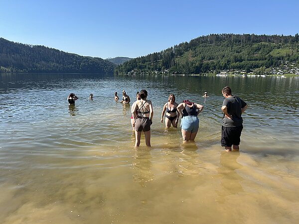 Eine Gruppe junger Menschen steht bis zu den Knien in einem Badesee; im Hintergrund sind bewaldete Hügel zu sehen.