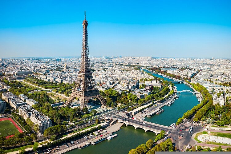 Neuer Partner aus Frankreich für den Bachelor-Top-Up Luftaufnahme von Paris mit dem Eiffelturm im Vordergrund, umgeben von Gebäuden, Parks und Straßen. Rechts im Bild fließt die Seine mit mehreren Brücken und Booten, der Himmel ist klar und blau.