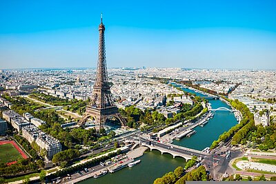 Luftaufnahme von Paris mit dem Eiffelturm im Vordergrund, umgeben von Gebäuden, Parks und Straßen. Rechts im Bild fließt die Seine mit mehreren Brücken und Booten, der Himmel ist klar und blau.