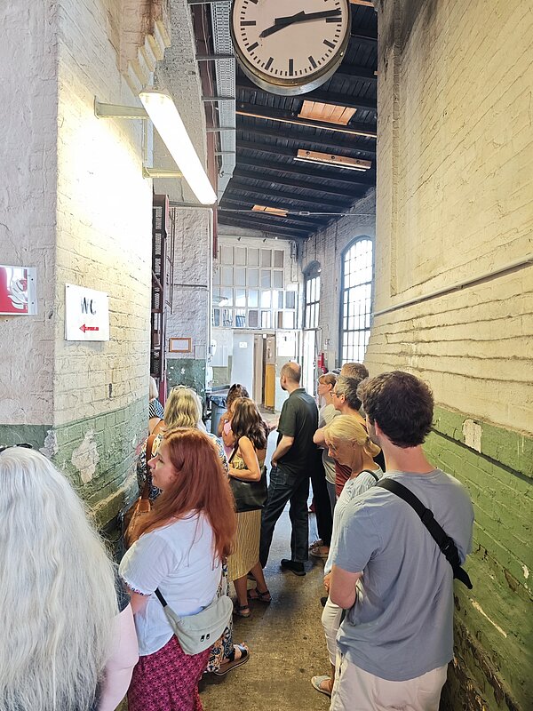 Eine Gruppe von Menschen sitzt an langen Holzbänken in einer großen, industriell gestalteten Halle. Die Halle ist mit Metallträgern und Oberlichtern ausgestattet. Im Hintergrund sind historische Züge und technische Geräte zu erkennen. Die Atmosphäre wirkt gesellig, und einige Personen unterhalten sich oder essen.