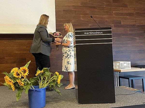 Eine Frau überreicht einer anderen Frau einen Blumenstrauß auf einer Bühne. Beide lächeln. Im Hintergrund ist ein Pult mit der Aufschrift "martinushaus" zu sehen. Auf dem Podium stehen Blumenarrangements mit Sonnenblumen.