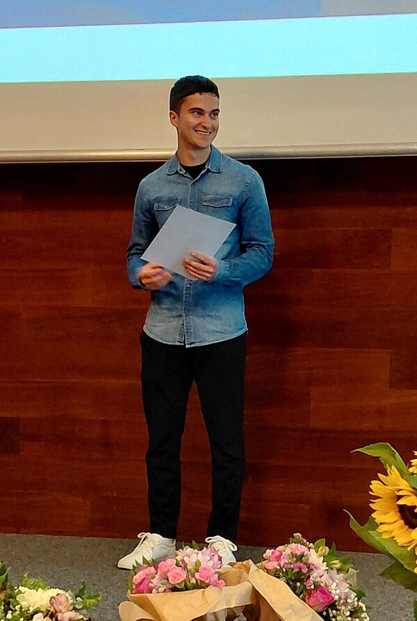 Ein junger Mann steht auf einer Bühne und hält ein Zertifikat. Er trägt ein blaues Hemd und lächelt. Auf dem Podium stehen Blumenarrangements mit Sonnenblumen.