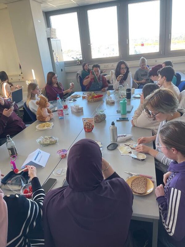 Eine Gruppe von Menschen sitzt in einem hellen Raum um einen großen Tisch, der mit Frühstückszutaten wie Brot, Aufstrichen, Obst und Getränken gedeckt ist. Einige Personen essen, andere unterhalten sich oder beschäftigen sich mit ihren Handys. Die Atmosphäre wirkt entspannt und gesellig. Im Hintergrund sind große Fenster mit Blick auf die Stadt zu sehen.