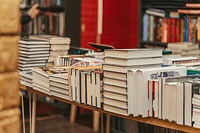Mehrere Bücherstapel sind ordentlich auf einem Holztisch aufgereiht. Im Hintergrund sieht man weitere Bücherregale, und eine rote Backsteinmauer ist teilweise zu erkennen. Die Szene vermittelt eine gemütliche und einladende Atmosphäre für Bücherliebhaber.