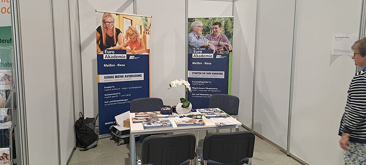 Messestand der Euro Akademie mit zwei Roll-Up-Bannern zu den Standorten Meißen und Riesa. Der Tisch in der Mitte ist mit Informationsmaterial und Dekoration ausgestattet. Zwei Stühle stehen bereit für Gespräche mit Interessierten. Eine Besucherin nähert sich dem Stand von rechts.
