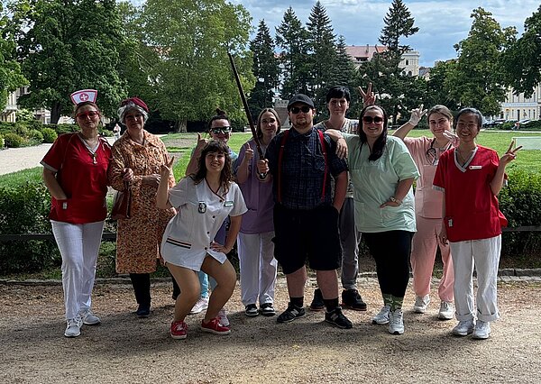 Die letzte Schulwoche der PFM 22: Ein unvergesslicher Abschluss Eine bunt gekleidete Gruppe von Personen steht draußen in einem Park. Viele sind als Pflegepersonal, Senior:innen oder in humorvollen Outfits verkleidet. Im Hintergrund ist eine Grünanlage mit Bäumen und Gebäuden zu sehen.