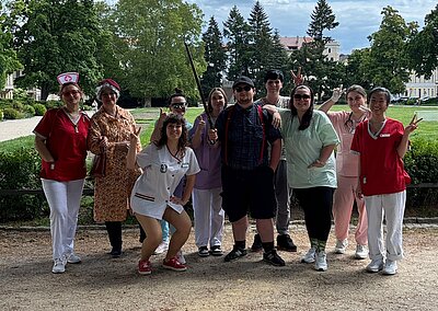 Eine bunt gekleidete Gruppe von Personen steht draußen in einem Park. Viele sind als Pflegepersonal, Senior:innen oder in humorvollen Outfits verkleidet. Im Hintergrund ist eine Grünanlage mit Bäumen und Gebäuden zu sehen.