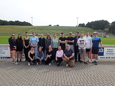 Eine große Gruppe junger Erwachsener posiert für ein Gruppenfoto auf einem Sportplatz. Im Hintergrund sind grüne Hügel und Sportanlagen zu sehen.