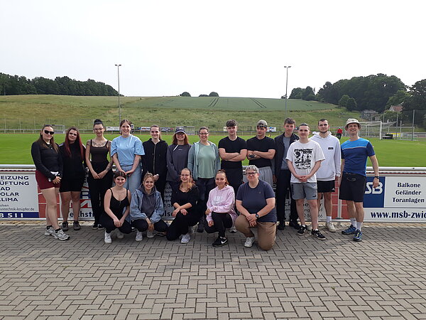 Unser Motto zum Sportfest: "Auf zu neuen Galaxien!" Eine große Gruppe junger Erwachsener posiert für ein Gruppenfoto auf einem Sportplatz. Im Hintergrund sind grüne Hügel und Sportanlagen zu sehen.