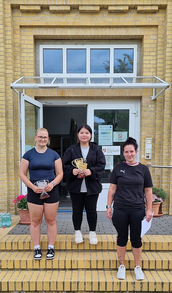 Wikinger-Sportfest: Pflegeazubis erobern die nordische Arena Zwei junge Frauen und eine weitere Frau stehen auf einer Treppe vor einem Gebäude. Sie halten Pokale und Urkunden in den Händen und posieren nach der Siegerehrung.