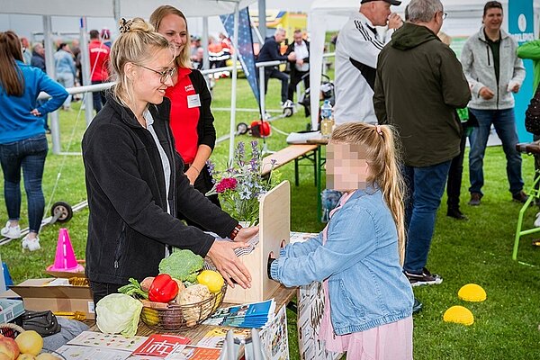 Benefiz Fußballspiel Auf einer Rasenfläche sind unter Zelten verschiedene Stände aufgebaut. An einem stehen zwei junge Frauen und reden mit einem kleinen Mädchen mit Zopf, das seine beiden Hände in eine Box steckt. Das Gesicht des Kindes ist verpixelt. Auf dem Tisch steht ein Korb mit Gemüse.