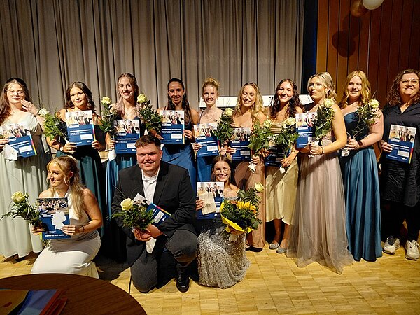 Absolventinnen und Absolventen posieren für ein gemeinsames Erinnerungsfoto. Alle halten ihre Abschlusszeugnisse sowie Blumen in Händen.