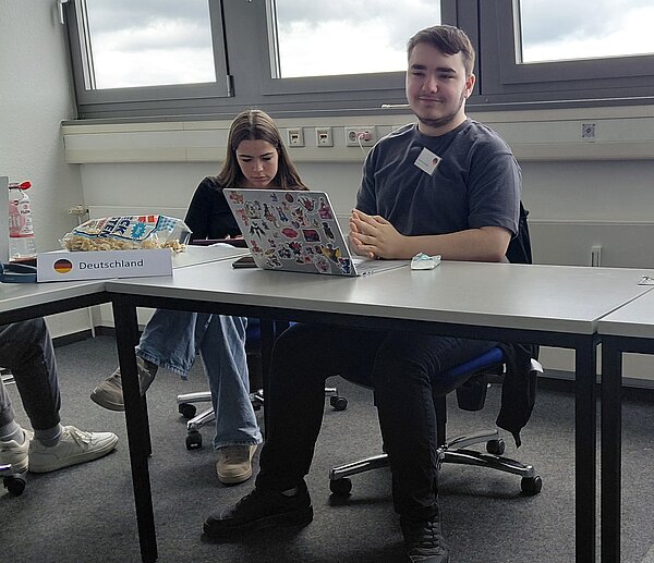 Zwei Schüler sitzen mit einem Laptop in einem Klassenzimmer, vor ihnen auf dem Tisch steht ein Schild mit der Aufschrift "Deutschland".