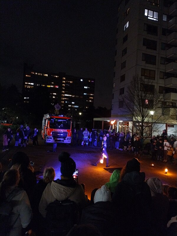 Große Menschenmenge schaut im Dunkeln einer Feuershow zu; vor einem Feuerwehrfahrzeug mit der Aufschrift „Feuerwehr“ jongliert eine Artistin mit brennenden Fackeln, ringsum hohe Wohnhäuser mit erleuchteten Fenstern.
