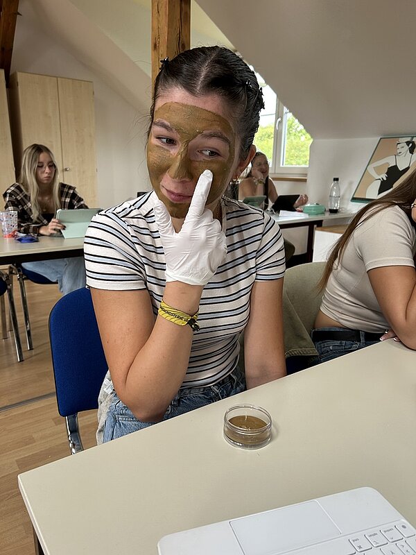 Eine weinbliche Auszubildende sitzt auf einem blauen Polsterstuhl am Tisch, trägt Einmalhandschuhe und sich selbst die Heilmaske aufs Gesicht auf.
