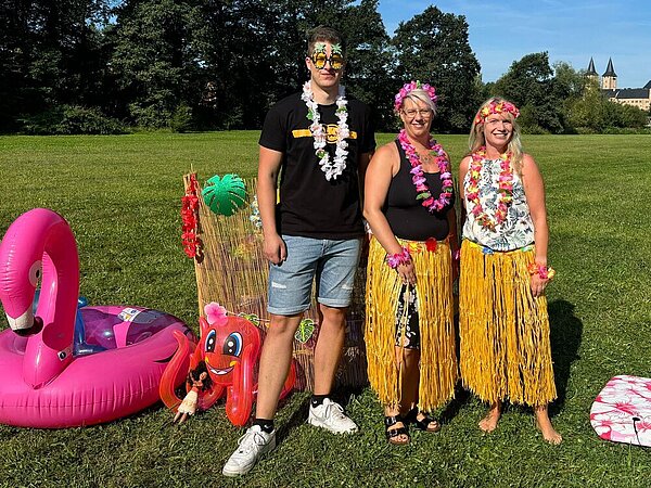 Drei Personen in Hawaii-Kostümen stehen auf einer Wiese. Neben ihnen liegen aufblasbare Wassertiere wie ein Flamingo und eine Krake. 