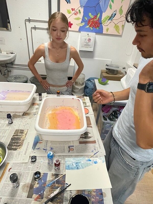 Zwei Personen arbeiten gemeinsam an einem Tisch. Eine Person mit weißem Tanktop und hochgestecktem Haar steht links und schaut in eine weiße Kunststoffwanne mit orange-rosa Farbverläufen. Die andere Person trägt graue Kleidung und eine Uhr am Handgelenk und schaut ebenfalls in die Wanne. Im Hintergrund sind ein Waschbecken und bunte Bilder an der Wand zu sehen.