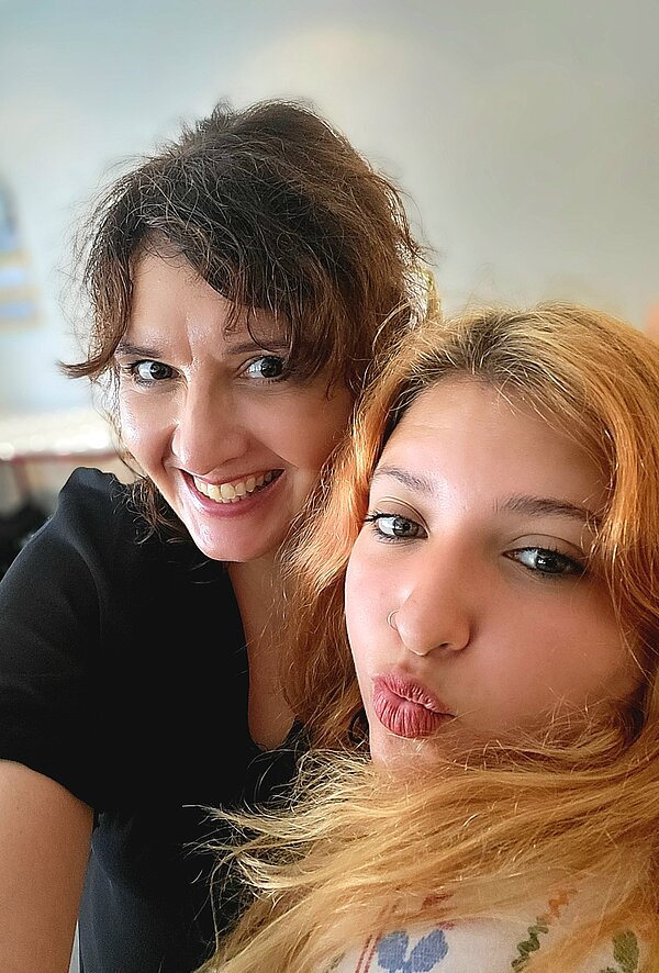 Zu sehen sind zwei lächelnde junge Frauen. Die linke Frau trägt ein schwarzes Oberteil und hat lockige braune Haare. Die rechte Frau hat ein weißes Oberteil an und hat hellblonde lockige Haare und macht einen Kussmund.