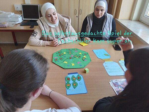 Vier Personen sitzen an einem Tisch und spielen ein selbstgestaltetes Brettspiel. Zwei Frauen mit Kopftuch sitzen auf der einen Seite des Tisches, während zwei andere Schülerinnen auf der gegenüberliegenden Seite sitzen und sich am Spiel beteiligen. Das Spielbrett ist grün mit bunten Blättern und Blüten, dazu gehören Spielkarten mit Zahlen und ein Würfel. Eine Person hebt die Hand, während die anderen lächeln oder konzentriert schauen. Der über das Bild gelegte Text lautet: „Das selbst ausgedachte Spiel der Fachschüler kam super an.“