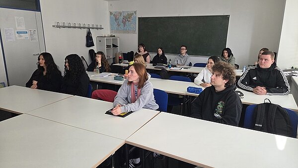 Ein Klassenzimmer zeigt die Auszubildenden der Euro Akademie Erfurt, die dem Hospizmitarbeiter zuhören, der über einen Vortrag hält. Die Azubis sitzen an weißen Tischen und schauen in Richtung des Mitarbeiters. An der Wand hängen eine Weltkarte und eine Tafel. Die Einrichtung wirkt schlicht, mit weißen Wänden und Decken. Die Personen tragen Alltagskleidung.