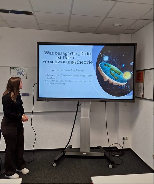 Eine Schülerin steht vor einem Bildschirm mit einer Folie zur „Flache-Erde“-Verschwörungstheorie. Die Darstellung zeigt eine grafisch illustrierte, flache Erde im Weltall.