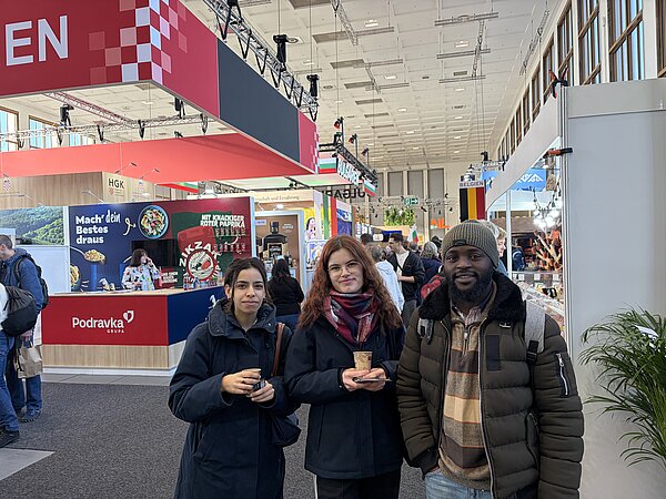 Drei Schüler stehen vor verschiedenen Ständen in einer Messehalle, zwei von ihnen halten Becher mit Getränken