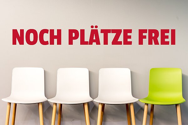 Eine Wand mit der Aufschrift „NOCH PLÄTZE FREI“ in roten Großbuchstaben. Darunter stehen drei weiße Stühle und ein grüner Stuhl, die an einer Wand aufgereiht sind.