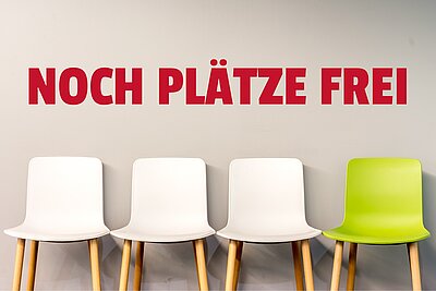 Eine Wand mit der Aufschrift „NOCH PLÄTZE FREI“ in roten Großbuchstaben. Darunter stehen drei weiße Stühle und ein grüner Stuhl, die an einer Wand aufgereiht sind.