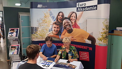 Infostand der Euro Akademie auf einer Messe. Zwei Frauen sitzen hinter einem Tisch mit Informationsmaterialien. Im Hintergrund ein großes Banner mit fröhlichen Jugendlichen und dem Schriftzug „Euro Akademie“. Auf dem Tisch liegen Flyer, Kugelschreiber und ein Schild mit der Standnummer 43.