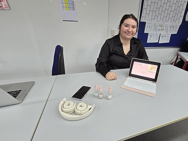 Eine Auszubildende mit dunkler, langärmeliger Bluse sitzt an einem Tisch. Vor ihr stehen ein Tablet mit hellem Tastaturcover, drei kleine Fläschchen mit rosafarbenem Deckel, ein Smartphone und ein rundes, cremefarbenes Kopfhöreretui mit Kopfhörern. Im Hintergrund sind ein Wandkalender, ein Lichtschalter und mehrere Informationsblätter an einer blauen Pinnwand zu sehen. Auf dem Nachbartisch steht ein aufgeklappter Laptop.