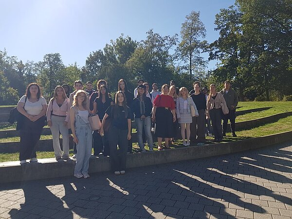 Gruppenfoto der Auszubildenden im Freien auf einer niedrigen Mauer vor einer Grünfläche mit Bäumen. Die Personen tragen unterschiedliche Freizeitkleidung, einige halten Taschen oder Rucksäcke. Die Sonne scheint, wodurch lange Schatten auf den Boden fallen. Im Hintergrund sind Bäume und blauer Himmel zu sehen.