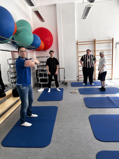 In einem hellen Raum mit weißen Wänden und großen Fenstern stehen vier Personen auf blauen Gymnastikmatten. Im Hintergrund befinden sich bunte Gymnastikbälle auf einem Regal sowie eine Sprossenwand. Die Personen tragen sportliche Freizeitkleidung und Socken. Eine Person steht mit verschränkten Armen auf einer Matte, die anderen drei stehen locker verteilt auf den übrigen Matten und schauen in seine Richtung. Im Raum liegen außerdem einige Handtücher und Sportgeräte.