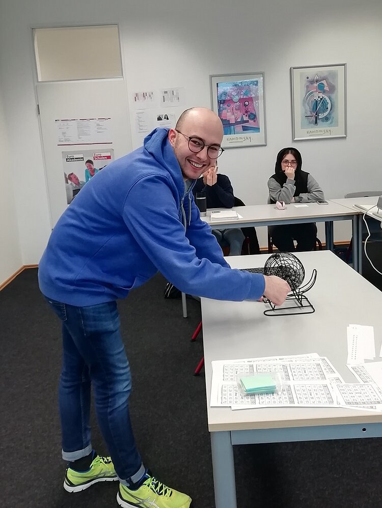 Mit Spaß im Spanisch Unterricht? – Bingo! Mitschüler in blauem Hoodie-Pullover dreht lächelnd an der Bingotrommel.