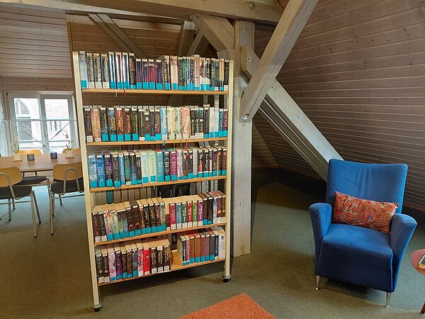 In einem Raum im Dachgeschoss mit holzverkleideten Wänden und Stützbalken aus Holz steht ein Regal voller Bücher, die mit blauen Aufklebern markiert sind. Neben dem Regal steht ein blauer Sessel mit einem Kissen. Im Hintergrund ist ein Tisch mit Stühlen und ein Fenster zu sehen.