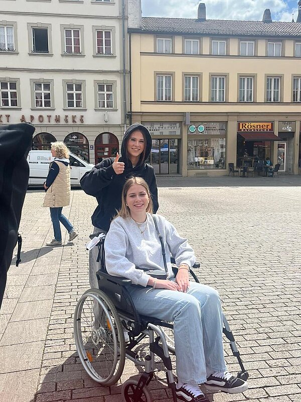 Protesttag zur Gleichstellung von Menschen mit Behinderung: Buttons für Inklusion Eine Schülerin sitzt im Rollstuhl, ihre Mitschülerin steht dahinter und macht einen Daumen hoch in Richtung Kamera.