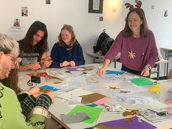 Eine Gruppe Studierender und eine Dozentin mit Rentiergeweih-Stirnband sitzen an einem Tisch und basteln weihnachtliche Anhänger aus buntem Papier und Prägefolie.
