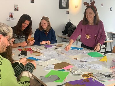 Eine Gruppe Studierender und eine Dozentin mit Rentiergeweih-Stirnband sitzen an einem Tisch und basteln weihnachtliche Anhänger aus buntem Papier und Prägefolie.