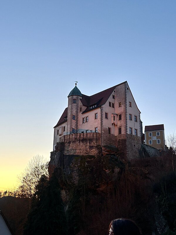 Historisches Schloss auf einem Felsen bei Sonnenuntergang, mit beleuchteten Fenstern und klarem Himmel in der Dämmerung.