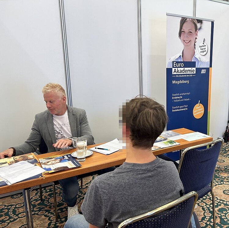 Ein Mitarbeiter der Euro Akademie Magdeburg mit grauem Sakko sitzt an einem Holztisch und spricht mit einem jungen Messebesucher. Auf dem Tisch liegen verschiedene Broschüren, ein Glas Wasser und ein Kugelschreiber. Im Hintergrund steht ein Roll-up mit dem Schriftzug 'Euro Akademie Magdeburg' und einem Porträt einer Frau. Der Raum hat helle Wände und einen gemusterten Teppichboden.