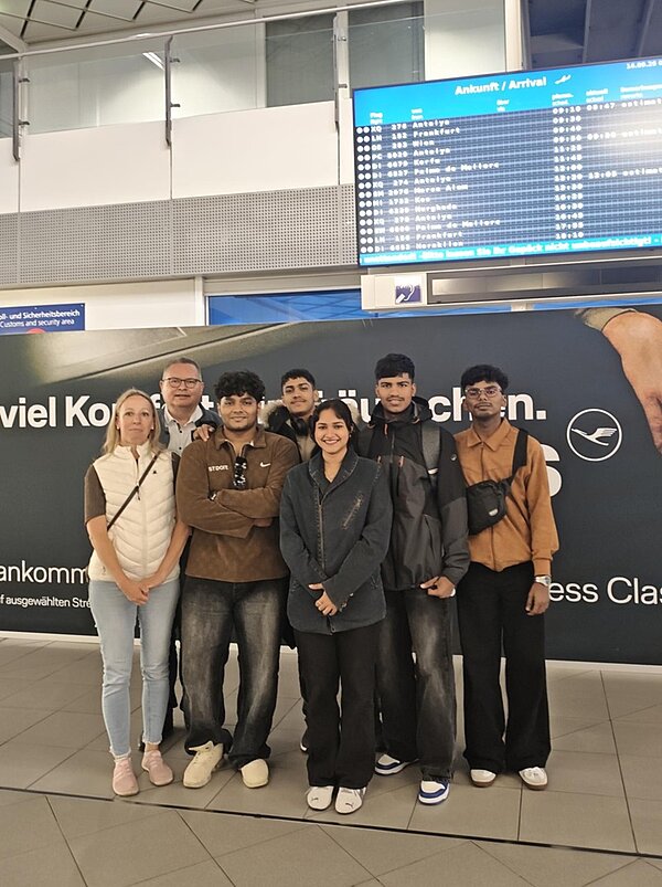 Eine Gruppe von Menschen steht in einem Flughafen vor einer großen Werbewand mit dem Logo einer Fluggesellschaft. Im Hintergrund befindet sich eine Anzeigetafel mit mehreren Zeilen, die Ankunftszeiten und Flugnummern aufführt. Die Personen tragen unterschiedliche Freizeitkleidung, darunter Jacken, Hemden und Hosen. Einige haben Taschen umgehängt. Die Stimmung wirkt entspannt, die Menschen stehen dicht beieinander und blicken in Richtung Kamera.