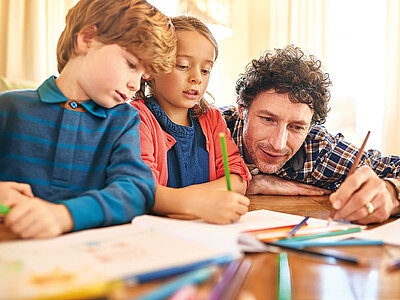 Ein Vater mit lockigen Haaren und einem karierten Hemd sitzt mit seinen beiden Kindern an einem Tisch und macht Hausaufgaben oder kreative Arbeiten. Der Junge links trägt ein blaues Langarmshirt mit Streifen und hält einen Stift in der Hand. Das Mädchen in der Mitte trägt ein dunkles Oberteil mit einer roten Jacke und malt konzentriert. Auf dem Tisch liegen viele bunte Stifte und Blätter. Alle drei wirken engagiert und konzentriert. Der Raum ist hell und freundlich.