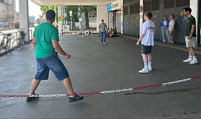 Eine Gruppe junger Menschen spielt unter einem überdachten Bereich vor einem Gebäude mit der Aufschrift 'Einfahrt'. Im Vordergrund steht ein Mann in grünem T-Shirt und blauer Shorts, der sich auf einen Ball oder ein Spiel konzentriert. Im Hintergrund sind weitere Personen zu sehen, die sich unterhalten oder zuschauen. Der Boden ist mit einer Kette abgesperrt.