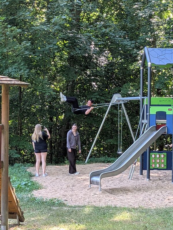 Eine Person schwingt auf einer Schaukel in einem Spielplatzbereich. Zwei weitere Personen stehen in der Nähe, eine davon hält ein Handy in der Hand und fotografiert die Szene. Im Hintergrund sind Bäume und im Vordergrund eine bunte Rutsche zu sehen.