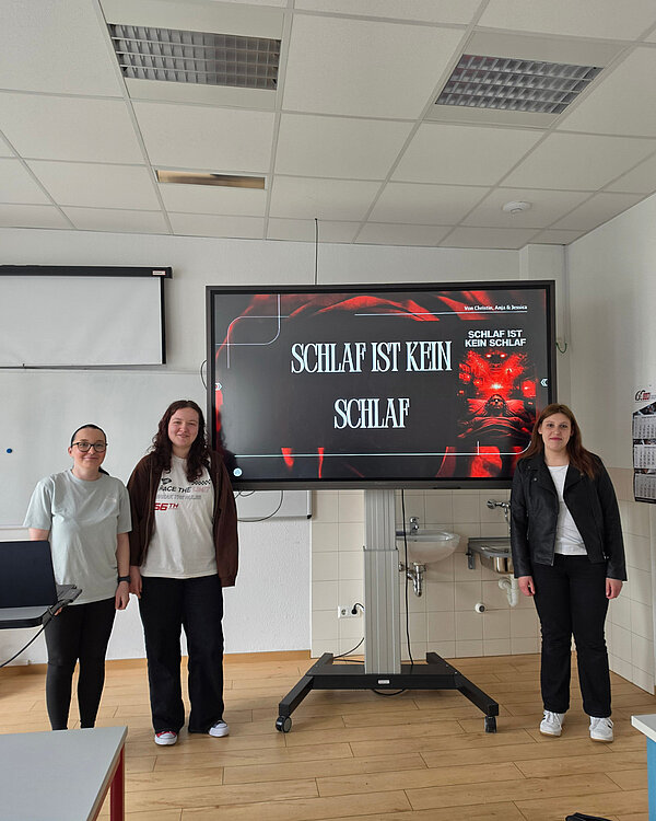 Drei Personen stehen vor einem Bildschirm, auf dem der Titel „Schlaf ist kein Schlaf“ von Christin, Anja und Jessica zu lesen ist. Die Folie zeigt eine düstere, rot beleuchtete Grafik.