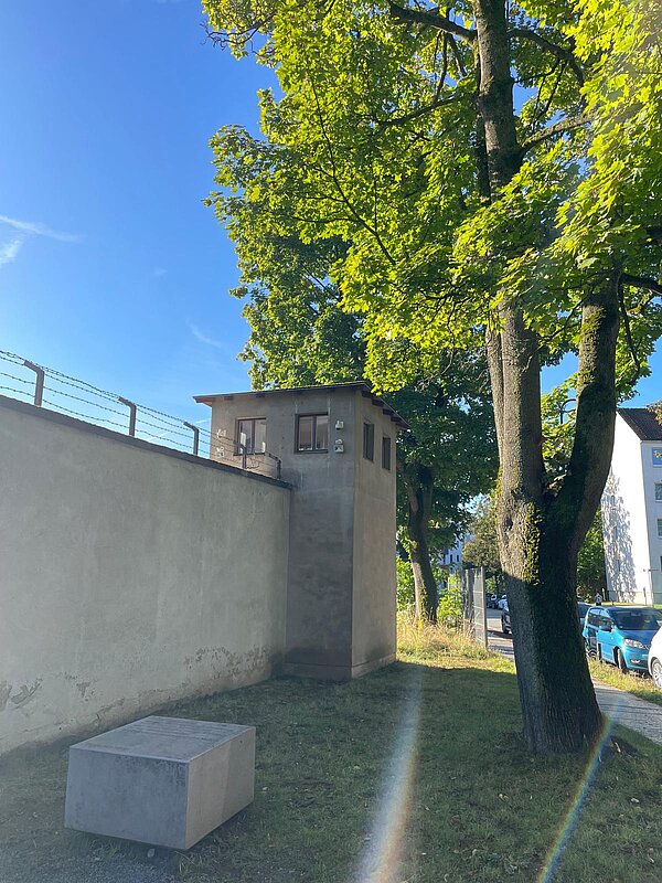 Ein ehemaliger Wachturm am Kaßberg-Gefängnis in Chemnitz, neben einer hohen Gefängnismauer mit Stacheldraht. Im Vordergrund steht ein Betonblock, daneben ein großer Baum unter blauem Himmel.