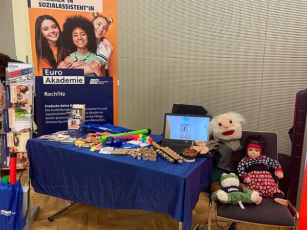 Blick auf den Messestand der Euro Akademie, mit großem Roll-Up und Tisch mit blauer Tischdecke, auf dem viele Infoflyer liegen und ein Stuhl mit Puppen.
