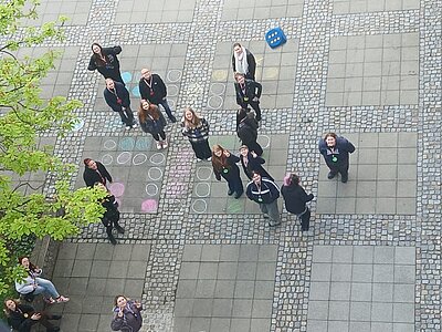 Auszubildende der Euro Akademie Dessau stehen auf einem gepflasterten Platz mit quadratischen Steinplatten und Kiesflächen. Einige blicken nach oben. Auf dem Boden sind bunte Kreidekreise und Zeichnungen zu sehen. Ein großer blauer Würfel liegt in einer Ecke. Links wächst ein Baum mit grünen Blättern.