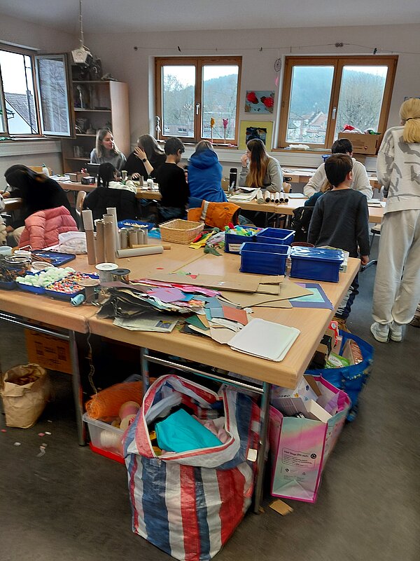 Großer Bastelraum mit vielen Personen an mehreren Tischen: Kinder und Jugendliche sitzen oder stehen, schneiden, kleben und gestalten, auf dem vorderen Tisch liegen zahlreiche bunte Papiere, Papprollen und Bastelmaterialien in Kisten und Taschen.